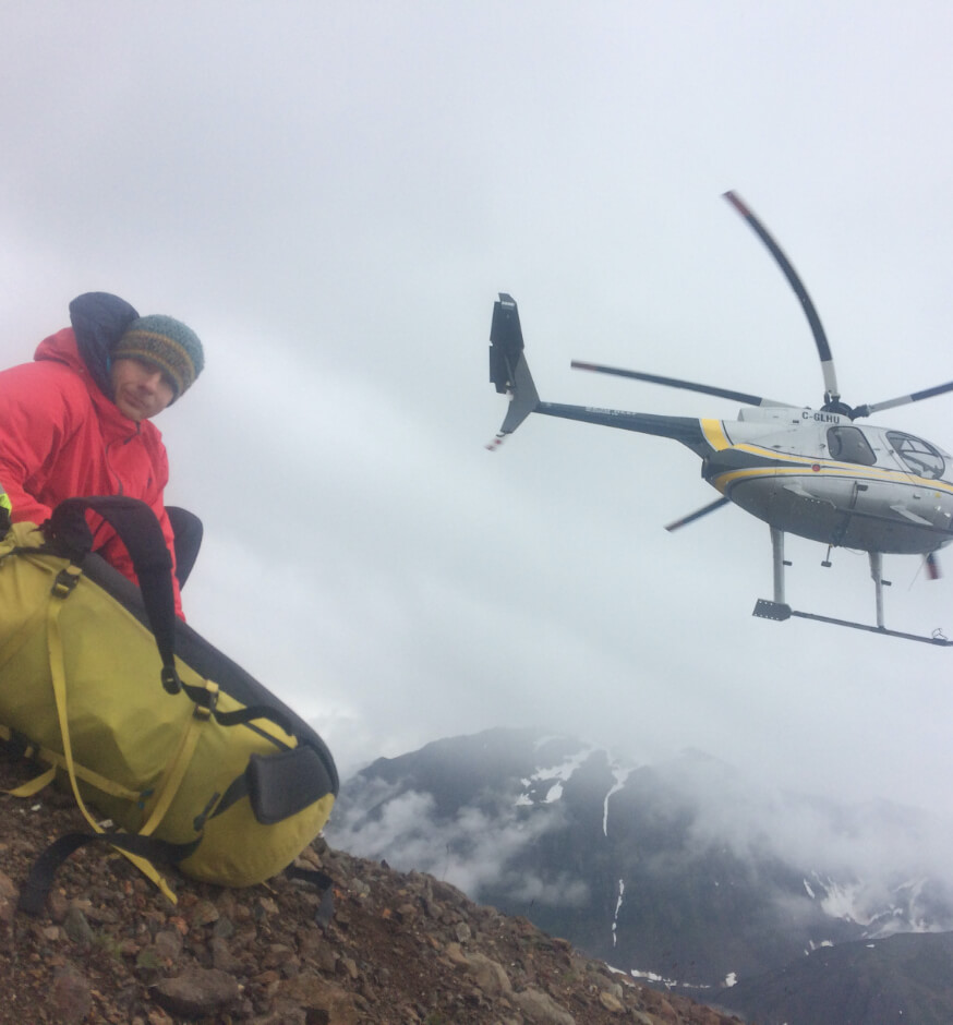 Man on Alaskan mountainside with gear waiting for helicopter to land.